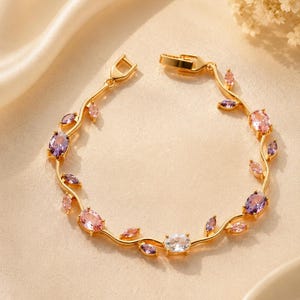 Op de afbeelding: Een goudkleurige armband met een fijne, rankachtige ketting, versierd met afwisselend ovale en marquise-geslepen edelstenen in tinten paars, roze en helder wit. Het sieraad is voorzien van een karabijnsluiting en een gepolijste afwerking.