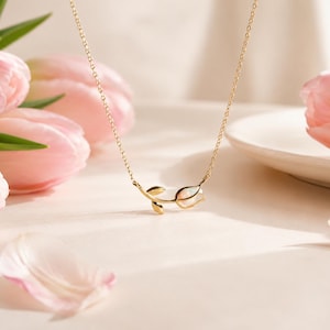 Puede incluir: Un collar dorado con un colgante de tulipán, sobre un fondo de tulipanes y pétalos rosas. La fina cadena y el colgante están enfocados, con una iluminación suave que realza el tema floral. El collar es una joya.