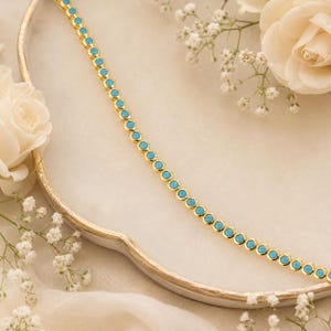 May include: A gold-tone chain necklace featuring a continuous row of small, round, bright turquoise-coloured bezel-set stones. The necklace is arranged in a diagonal line across the frame, showcasing a uniform design with polished metal edges.