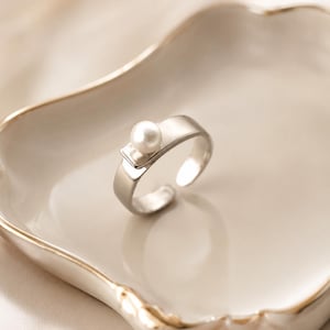 Könnte beinhalten: Ein silberfarbener Ring mit offener Ringschiene, der mit einer einzelnen, glatten, runden weißen Perle auf einer kleinen rechteckigen Plattform besetzt ist. Der Ring hat eine polierte Metalloberfläche und eine Öffnung zur Größenanpassung.