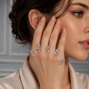 May include: A silver-toned hand ornament featuring four delicate leaf-shaped motifs adorned with small, clear, sparkling stones. Each leaf design is connected by a thin metal band, creating a continuous piece that rests across the fingers.