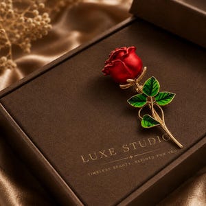 May include: A rose brooch featuring a vibrant red flower head with a gold-toned stem and bright green leaves. The brooch is placed on a dark brown rectangular box with the text LUXE STUDIO and TIMELESS BEAUTY, REFINED FOR YOU printed in gold.