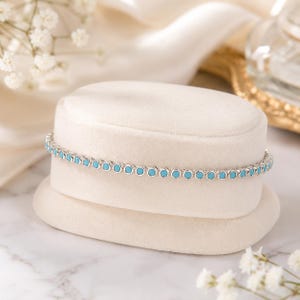May include: A silver-toned tennis bracelet featuring a continuous row of small, round, bright turquoise-coloured stones set in individual metal bezels, creating a uniform and linked chain design that wraps around a cream-coloured display stand.