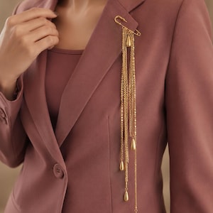 May include: A gold-toned safety pin brooch featuring multiple dangling chains of varying lengths. Each chain terminates in a small, smooth, teardrop-shaped charm. The brooch is pinned to the lapel of a solid mauve-colored blazer worn over a matching top.