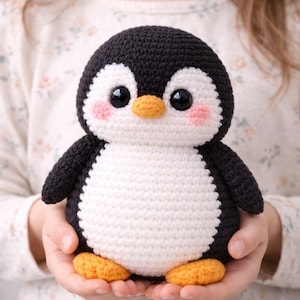 Op de afbeelding: Een handgemaakt gehaakt pinguïn knuffel. De pinguïn heeft een zwarte kop en vleugels, een witte buik en oranje voeten en snavel. De wangen van de pinguïn zijn roze. Het speelgoed wordt in de handen van een persoon gehouden.