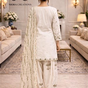 Puede incluir: Conjunto salwar kameez blanco con bordados intrincados. El atuendo incluye una t&uacute;nica, pantalones sueltos y una bufanda a juego. El bordado presenta patrones florales y de hojas en un color complementario. De Niora Creations.