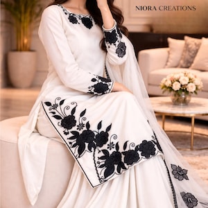 Puede incluir: Conjunto blanco con intrincado bordado floral negro. El atuendo incluye una parte superior de manga larga y pantalones de pierna ancha, con detalles florales en el escote, las mangas y el dobladillo. Una bufanda transparente a juego completa el look. La marca "NIORA CREATIONS" es visible.