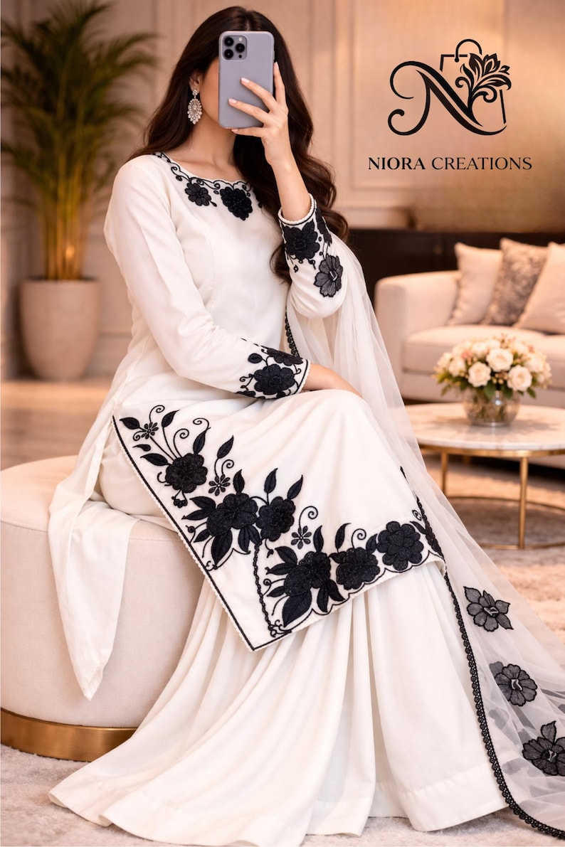 Puede incluir: Conjunto blanco con intrincado bordado floral negro. El atuendo incluye una parte superior de manga larga y pantalones de pierna ancha, con detalles florales en el escote, las mangas y el dobladillo. Una bufanda transparente a juego completa el look. La marca "NIORA CREATIONS" es visible.
