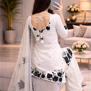Puede incluir: Traje salwar kameez blanco con bordado floral negro. El conjunto incluye una t&uacute;nica de manga larga, pantalones de pernera ancha y un dupatta transparente. La parte trasera de la t&uacute;nica presenta un detalle de lazo y borlas. La marca "NIORA CREATIONS" es visible.