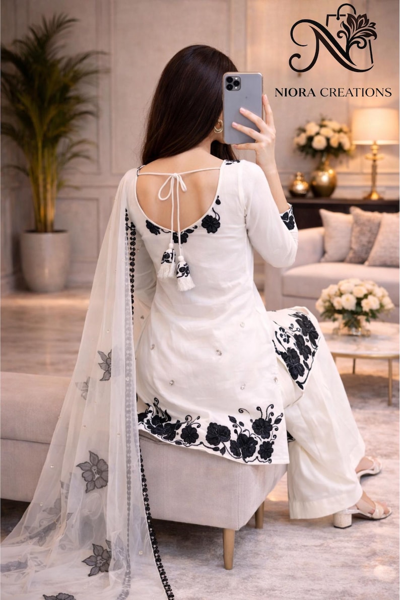 Puede incluir: Traje salwar kameez blanco con bordado floral negro. El conjunto incluye una t&uacute;nica de manga larga, pantalones de pernera ancha y un dupatta transparente. La parte trasera de la t&uacute;nica presenta un detalle de lazo y borlas. La marca "NIORA CREATIONS" es visible.