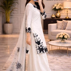 Puede incluir: Pantalones palazzo blancos y t&uacute;nica a juego de manga larga con bordados florales negros. Una bufanda blanca transparente con detalles florales negros cubre el conjunto. El conjunto es de Niora Creations.