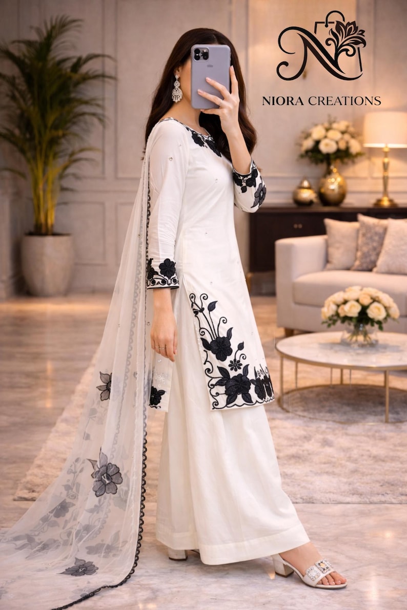 Puede incluir: Pantalones palazzo blancos y t&uacute;nica a juego de manga larga con bordados florales negros. Una bufanda blanca transparente con detalles florales negros cubre el conjunto. El conjunto es de Niora Creations.