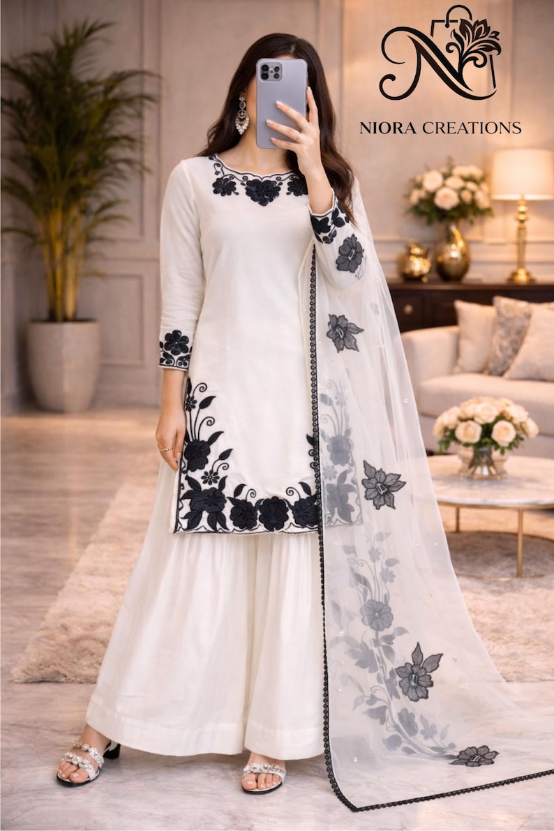 Puede incluir: Conjunto blanco con t&uacute;nica de manga larga y pantal&oacute;n de pernera ancha, adornado con bordados florales negros. Una bufanda transparente a juego con detalles florales completa el atuendo. El nombre de la marca "NIORA CREATIONS" es visible.