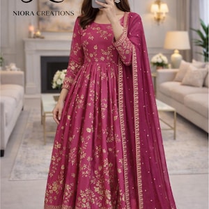 May include: A magenta-colored, long-sleeved dress with gold floral embroidery, matching pants, and a dupatta. The dress features a round neckline and a flared skirt. The image includes the text "NIORA CREATIONS".