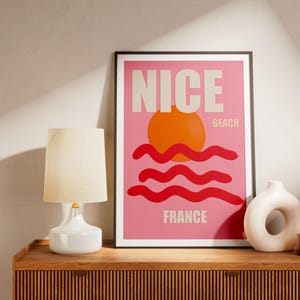 May include: A framed poster with the word "NICE" in large, off-white letters, above a stylized orange sun and red waves on a pink background. The word "FRANCE" is at the bottom. A white lamp and vase are on a wooden surface.