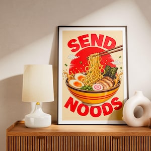 Peut inclure: Impression d'art encadrée représentant un bol de ramen avec des baguettes, un soleil rouge et les mots "SEND NOODS" en rouge. L'œuvre est exposée sur un meuble en bois, à côté d'une lampe blanche et d'un vase décoratif.