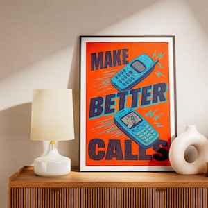 Peut inclure: Impression d'art encadrée avec un fond orange vif et les mots "MAKE BETTER CALLS" en bleu. Deux téléphones portables bleus rétro sont illustrés. Une lampe et un vase blancs sont sur une surface en bois.