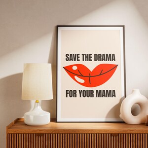 May include: A framed art print with the text "SAVE THE DRAMA FOR YOUR MAMA" above a red lip illustration. The print is displayed on a wooden cabinet next to a white lamp and a decorative vase.