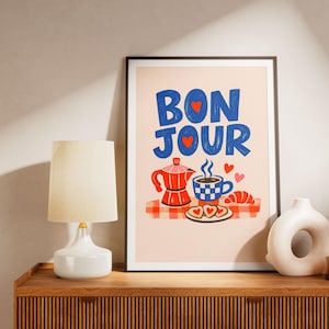 May include: A framed art print with the word "BONJOUR" in blue letters with red hearts. The print features a red coffee pot, a checkered cup of coffee, a croissant, and heart-shaped cookies. A white lamp and vase are on a wooden cabinet.