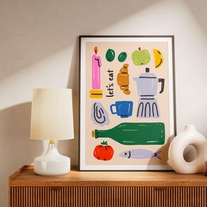 Pode incluir: Uma impressão artística emoldurada com ilustrações coloridas de alimentos, incluindo um croissant, uma maçã, uma cafeteira e uma garrafa. As palavras "let's eat" estão escritas na impressão. A impressão é exibida em uma superfície de madeira com uma lâmpada e um vaso.