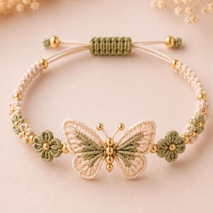 May include: A macrame bracelet featuring a central butterfly motif in cream and sage green cord, accented with small gold-toned beads. The design includes two flower-shaped knots on either side and an adjustable sliding closure with gold-toned beads.