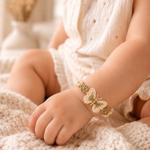 A baby's arm wearing a beige and green crocheted butterfly bracelet with golden beads, resting on a textured cream blanket.
