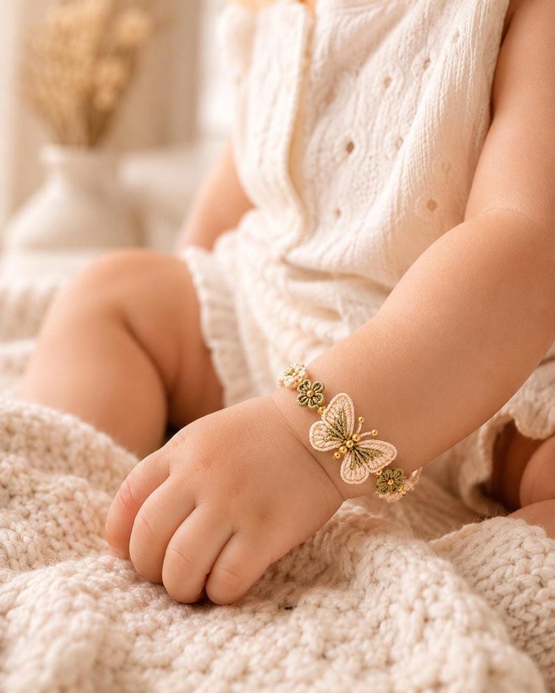 A baby's arm wearing a beige and green crocheted butterfly bracelet with golden beads, resting on a textured cream blanket.