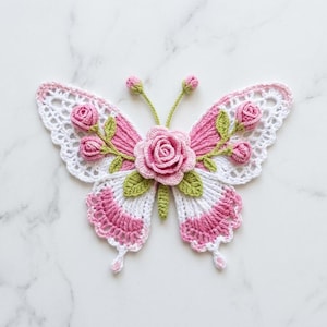 May include: A crochet butterfly applique featuring a central pink rose, green leaves, and small pink rosebuds. The wings are crafted with white and pink yarn in a delicate, openwork pattern. Two thin green antennae with small buds extend upward.