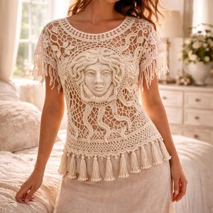 Puede incluir: Top de crochet color crema con un diseño de rostro de Medusa en la parte delantera. El top presenta mangas cortas con flecos y un ribete de borlas en la cintura. El top se combina con una falda beige.