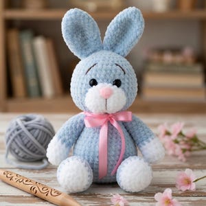 May include: A light blue crochet bunny with white accents on the paws and ears, and a pink bow. The bunny has black eyes and a pink nose. A ball of gray yarn and a wooden crochet hook are next to the bunny.