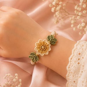 A hand wearing a beige and green crocheted bracelet with a central yellow flower and green leaves, on a pink satin fabric.