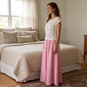 A woman with long brown hair stands in a bedroom, wearing a white short-sleeved crochet top with a floral openwork pattern and a long pink skirt.