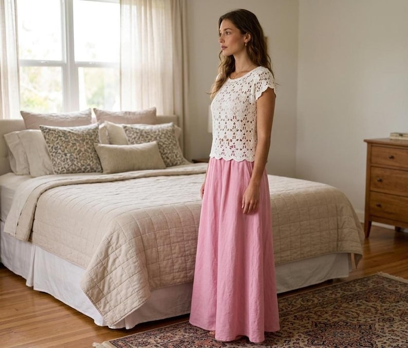 A woman with long brown hair stands in a bedroom, wearing a white short-sleeved crochet top with a floral openwork pattern and a long pink skirt.