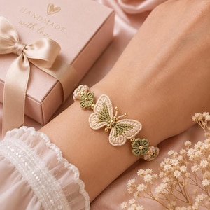A hand wearing a beige and green crocheted butterfly bracelet with golden beads, next to a pink gift box with a ribbon.