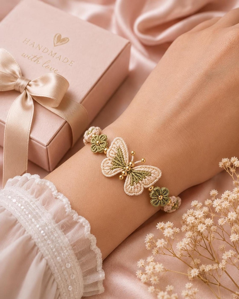 A hand wearing a beige and green crocheted butterfly bracelet with golden beads, next to a pink gift box with a ribbon.
