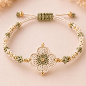 A beige and green crocheted bracelet featuring a central four-petal flower design with golden beads, on a light pink surface.