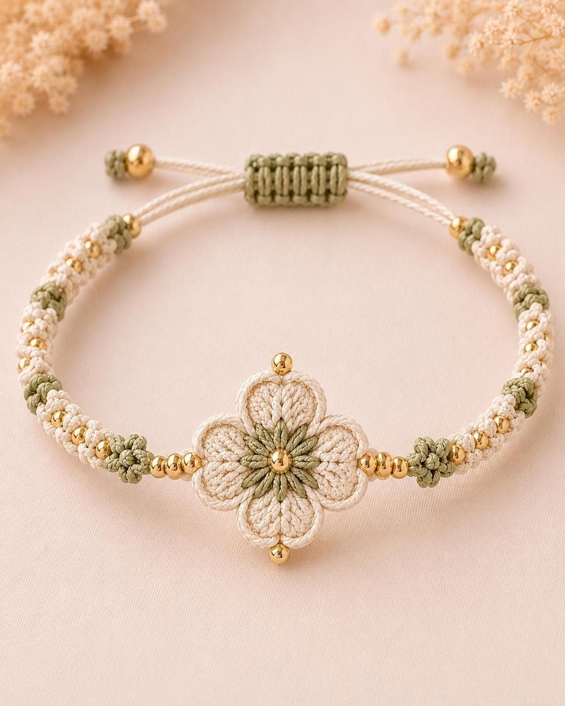 A beige and green crocheted bracelet featuring a central four-petal flower design with golden beads, on a light pink surface.