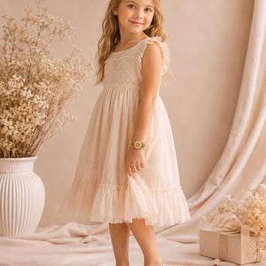 A young girl in a cream dress with ruffled sleeves stands, wearing a yellow crocheted flower bracelet on her right wrist.