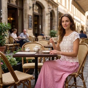 A woman with long brown hair sits at an outdoor cafe table, wearing a white short-sleeved crochet top with a floral openwork pattern and a long pink skirt, holding a teacup.
