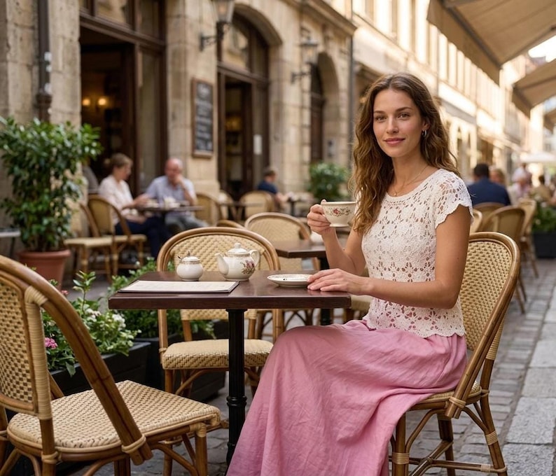 A woman with long brown hair sits at an outdoor cafe table, wearing a white short-sleeved crochet top with a floral openwork pattern and a long pink skirt, holding a teacup.