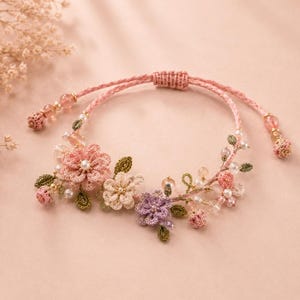 Op de afbeelding: Een delicate armband met gehaakte bloemen in roze, crème en paarse tinten, geaccentueerd met kleine groene blaadjes, witte parels en doorschijnende kralen. Het sieraad is gemaakt met een roze koord en een verstelbare schuifknoopsluiting.