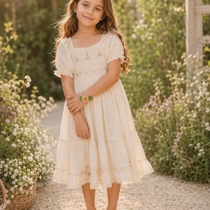 A young girl in a cream dress with short sleeves and embroidered bodice stands outdoors, wearing a yellow crocheted flower bracelet on her left wrist.