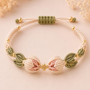 Könnte beinhalten: Ein Makramee-Armband mit zwei zentralen Blütenknospen in Creme und zartem Rosa, flankiert von olivgrünen, blattförmigen Knoten. Das Design enthält kleine goldfarbene Metallperlen und einen verstellbaren Schiebeknotenverschluss aus cremefarbener Kordel.