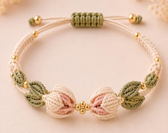 May include: A macrame bracelet featuring two central flower buds in cream and soft pink, flanked by olive green leaf-shaped knots. The design includes small gold-toned metallic beads and an adjustable sliding knot closure with cream-coloured cord.
