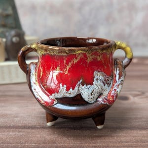 May include: A small ceramic pot with a glossy glaze. The pot has a vibrant red interior with a dripped effect, transitioning to brown and yellow at the rim. It has two handles and three small feet, with white accents on the base.