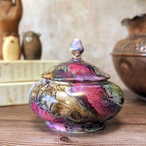 May include: A small, rounded ceramic lidded jar featuring a vibrant, swirling abstract pattern in shades of magenta, deep gold, and dark brown. The lid is topped with a pointed finial, and the entire surface has a glossy, reflective finish.