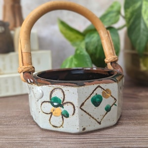 May include: A hexagonal ceramic basket with a speckled off-white glaze, featuring hand-painted green and gold floral and geometric motifs. The vessel includes a curved, light brown bamboo handle attached to the sides with woven rattan accents.