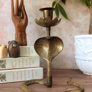 May include: A brass candlestick holder shaped like a cobra snake with its hood flared. The snake's body forms the base, and a lotus-shaped cup sits atop the cobra's head. The metal surface features intricate engraved leaf and scale patterns.