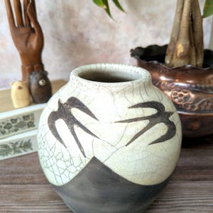 May include: A round ceramic vase with a cream-coloured upper section featuring a dark crackle glaze pattern and two black bird silhouettes. The lower portion of the vase is a solid matte charcoal grey. The vase has a wide, circular opening at the top.