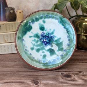 May include: A round ceramic bowl with a brown rim and a white interior featuring a mottled pattern of green and deep blue glaze. The centre of the bowl displays a concentrated cluster of dark blue and white spots, creating a textured appearance.